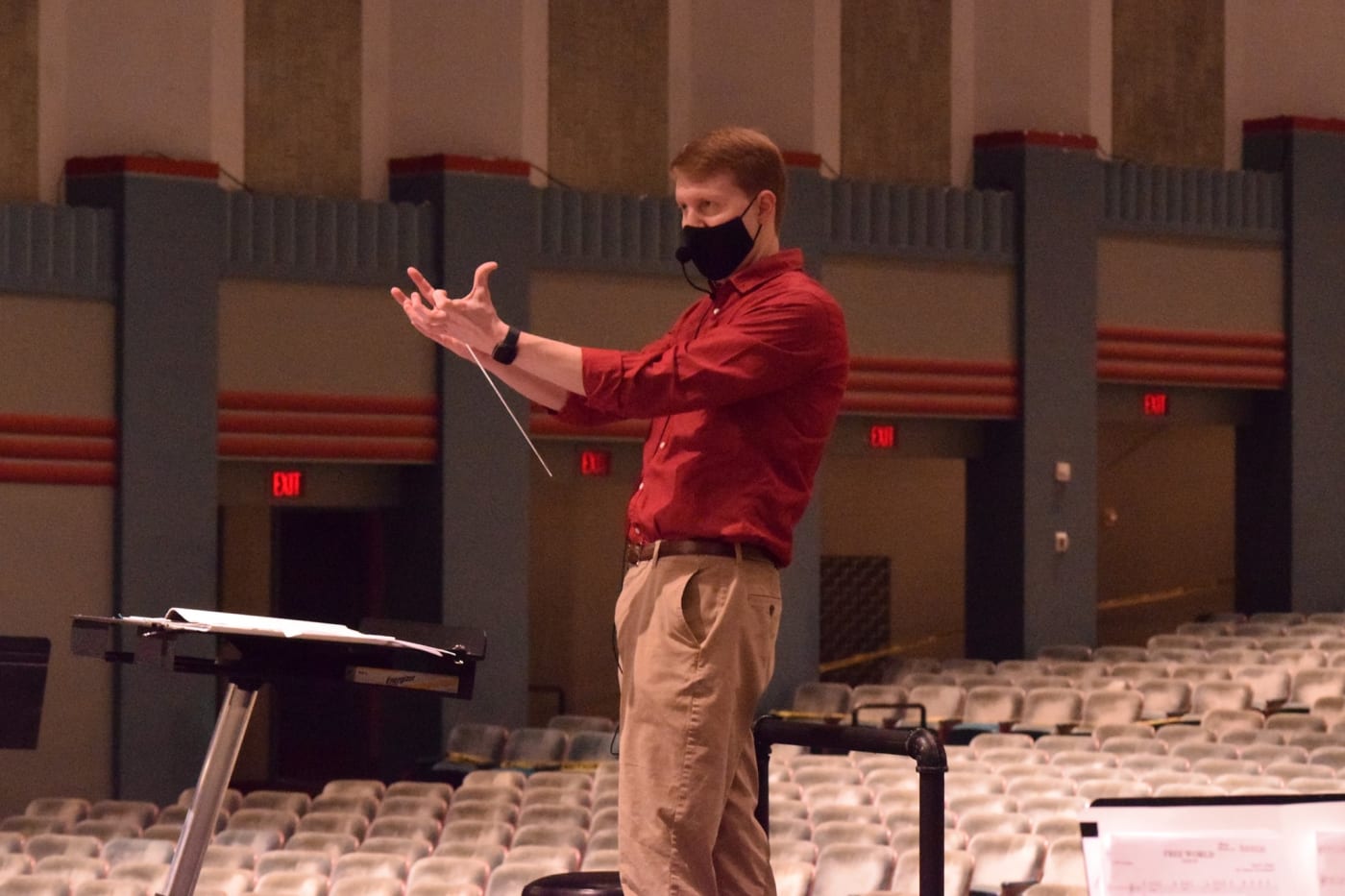 A man stands on a conducting podium, baton flipped between fingers. His hand are directly in front of his chest as he draws sound out from the group in front of him.